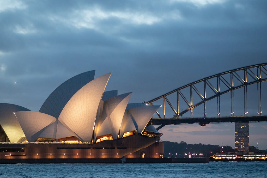 Facade of modern Sydney Opera House of unusual design with large concrete shells located on river coast near massive bridge at night